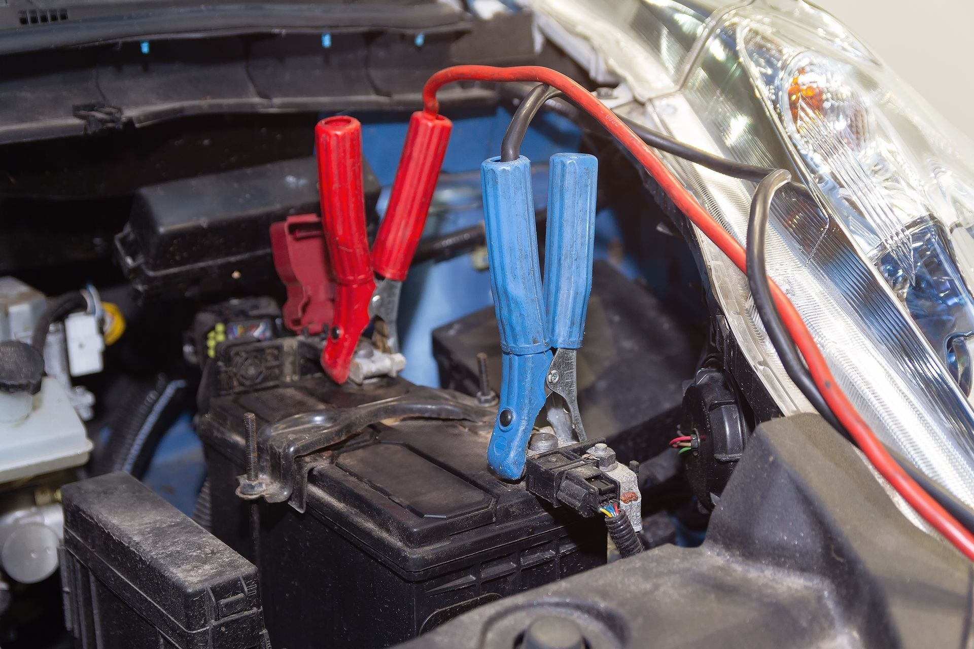 Car battery in engine bay with red and blue jumper cable clamps attached to the terminals