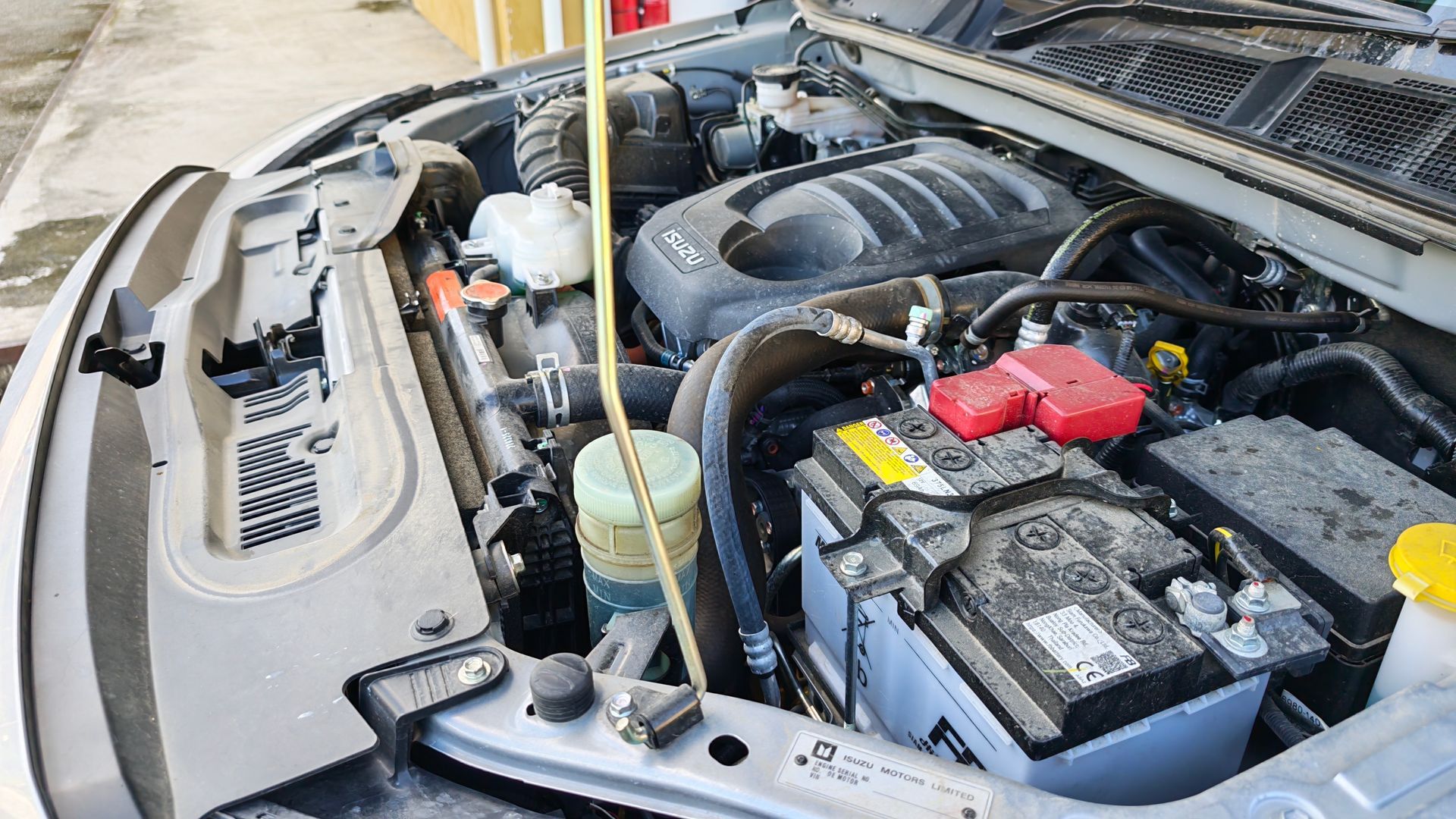Car engine bay with open hood, battery, coolant reservoir, and hoses visible.