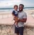 Man holding a smiling child on a seaside promenade with ocean waves in the background