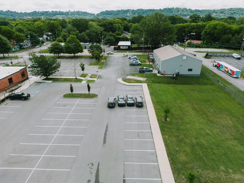 Empty parking lot beside a white building and grassy field, with trees in the background.