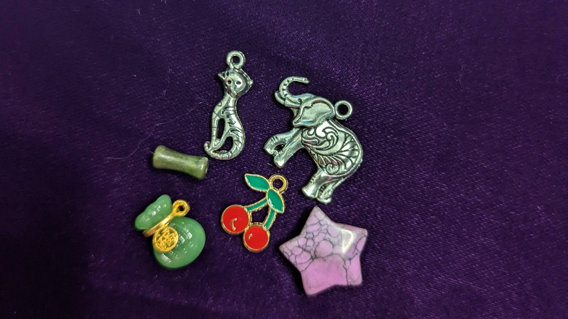 Assorted small jewelry charms on purple fabric, including silver, green, red cherry, and pink star shapes