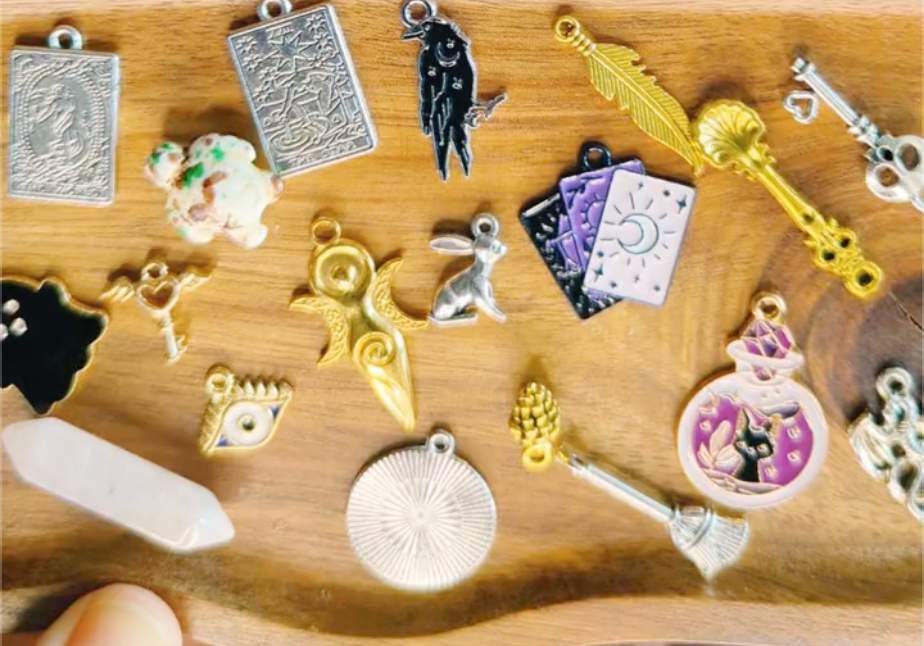 Collection of colorful charms on a wooden tray including broom, key, feather, crow, tarot card, ghost, crystal, and more