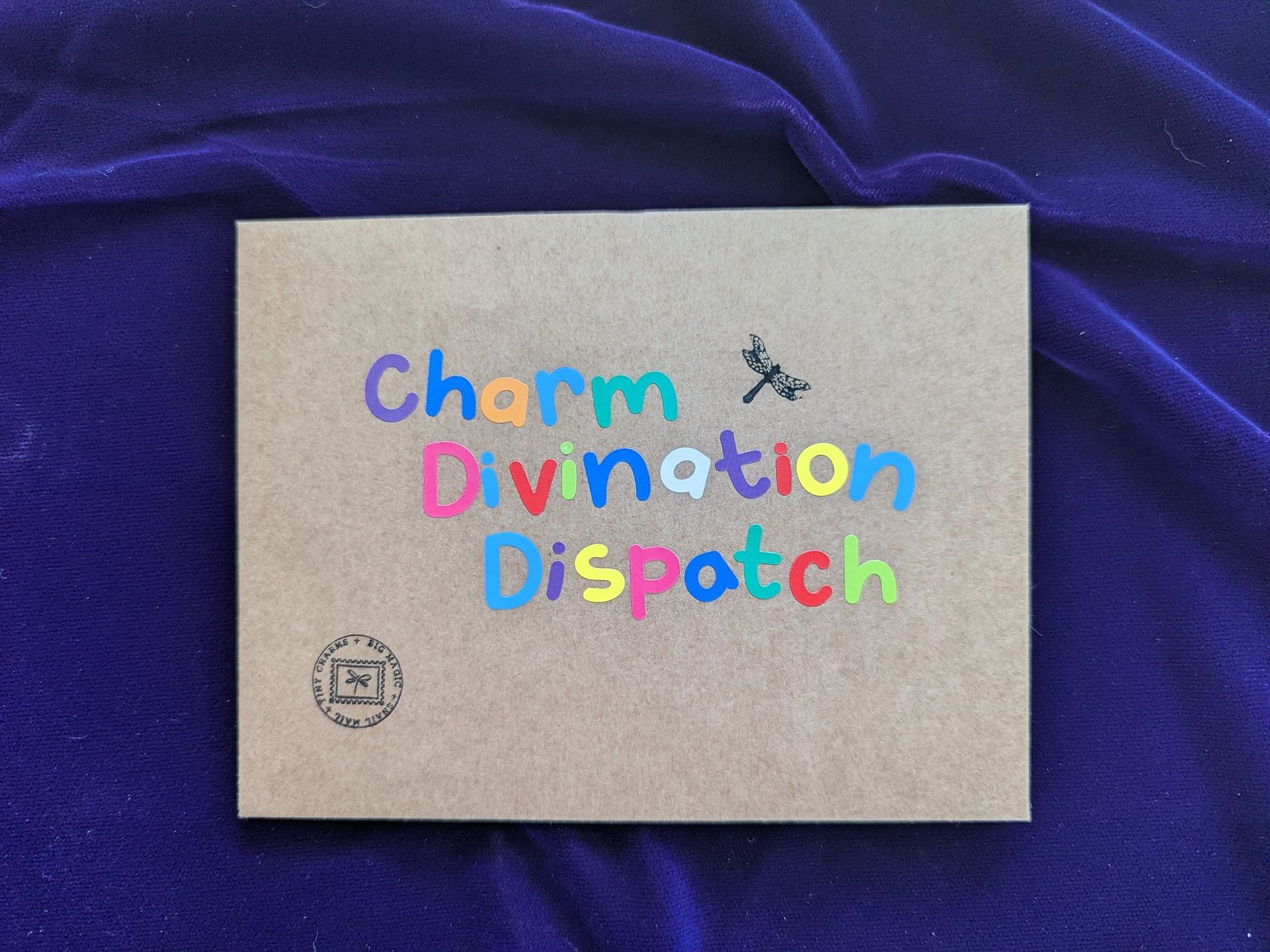 Colorful “Charm Divination Dispatch” text on a small beige card against purple fabric.