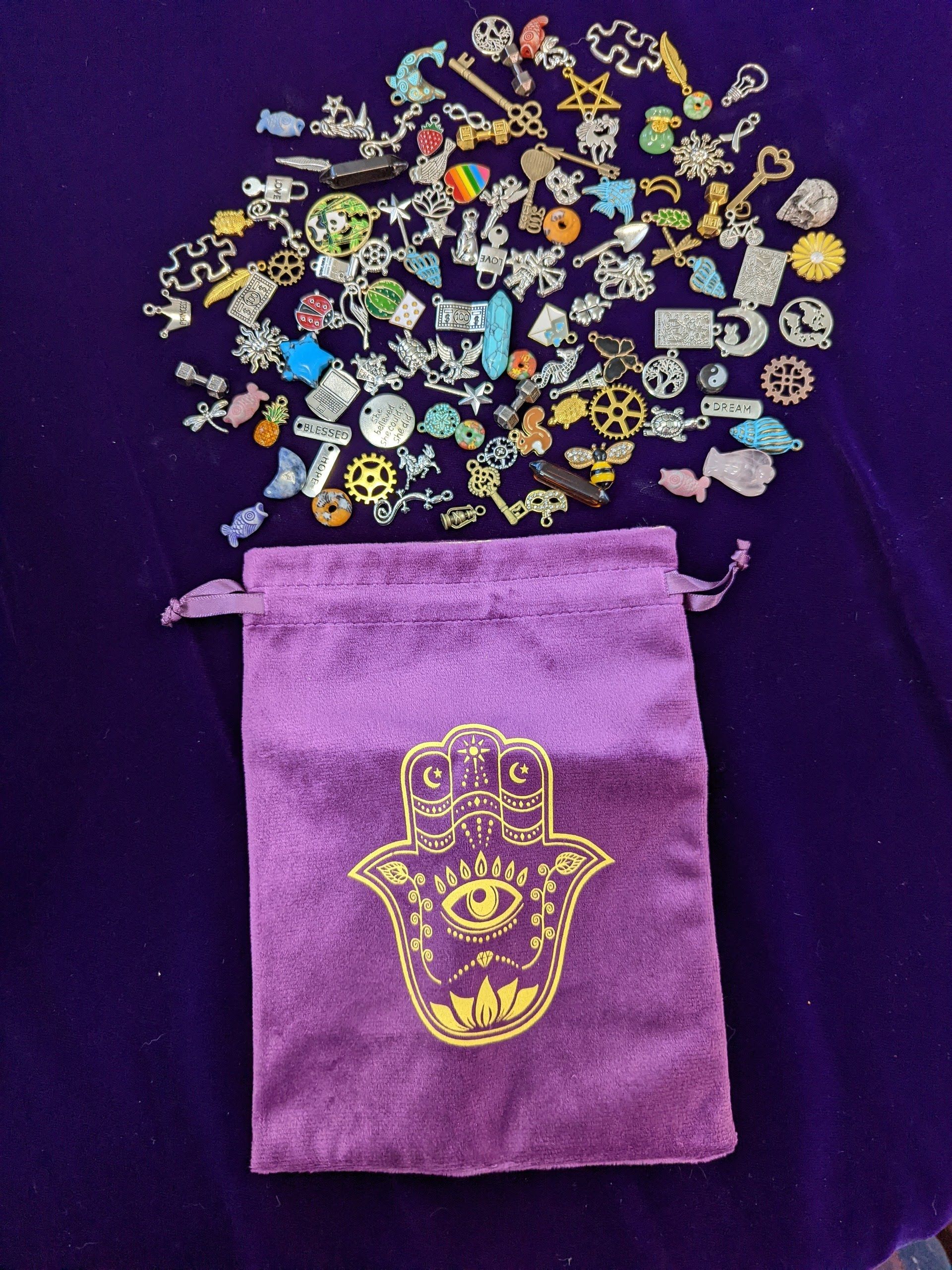 Purple drawstring pouch with gold hamsa emblem, spilled open to colorful small charms above it