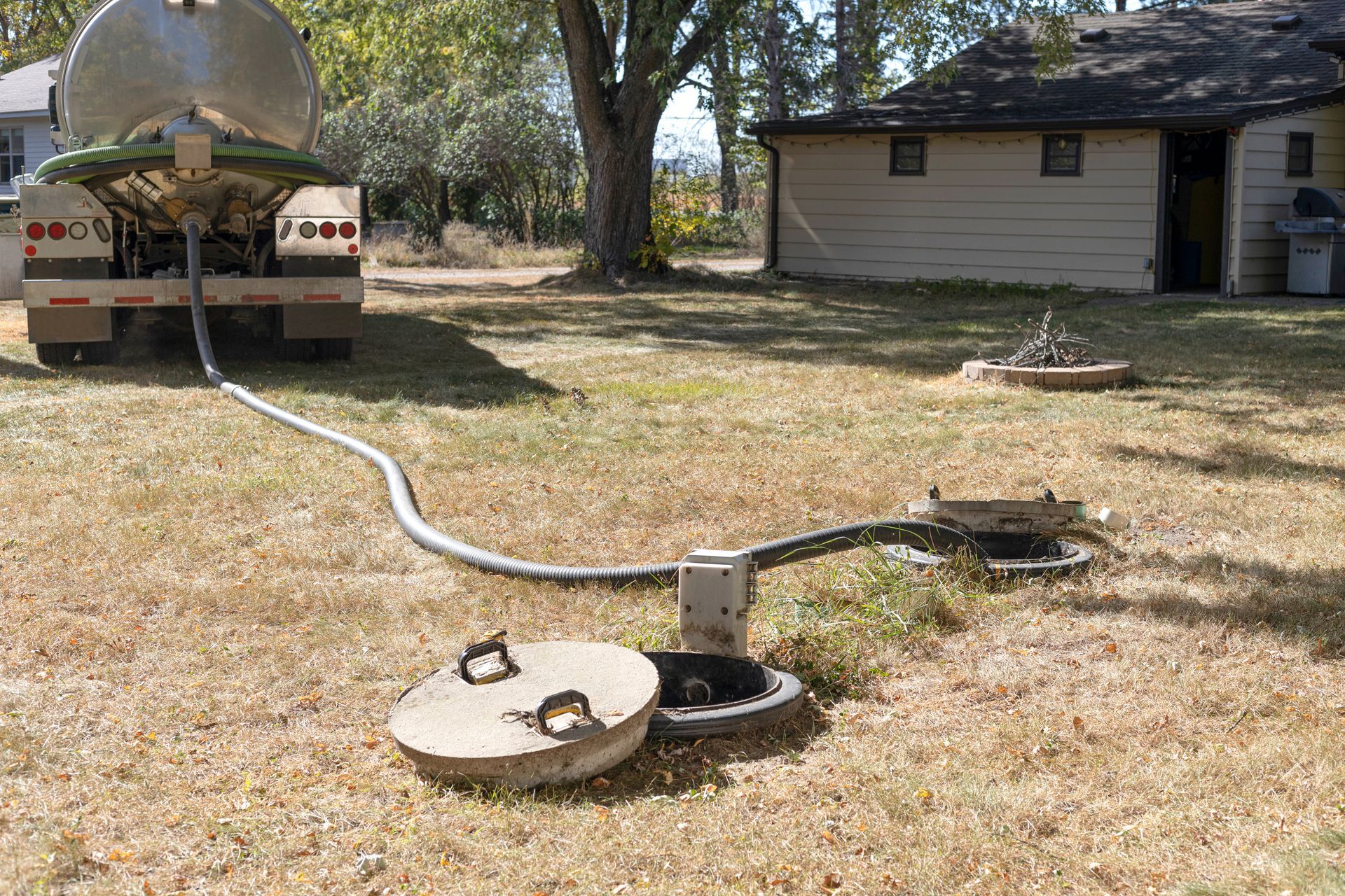 Sealing a pre-cast concrete sewage manhole with mortar during a new septic tank installation. Rural yard with two utility access covers connected by a hose or pipe, beside a house and truck trailer.