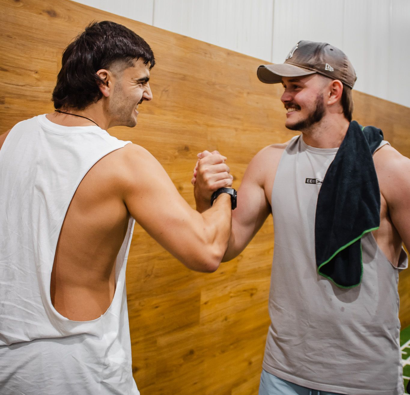 Two people in gym tanks perform a handshake in front of a wooden wall.