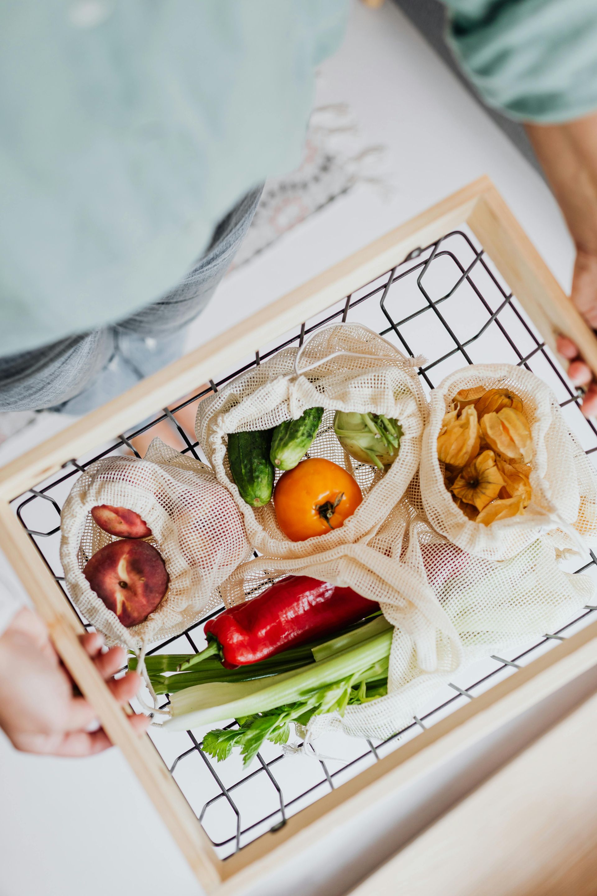 A person holds a wooden tray containing fresh produce in reusable mesh produce bags, including vegetables and fruit.