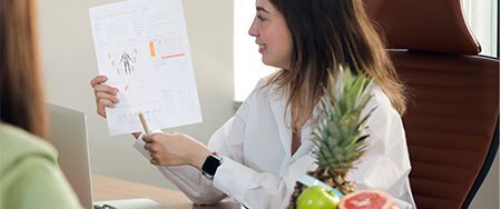 Woman pointing at a chart while sitting at a desk with fruit, possibly a nutritionist.