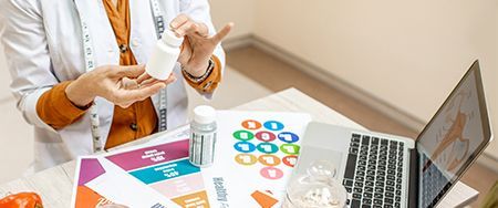 Person in lab coat holding a pill bottle, possibly a healthcare consultation. Laptop and papers on table.