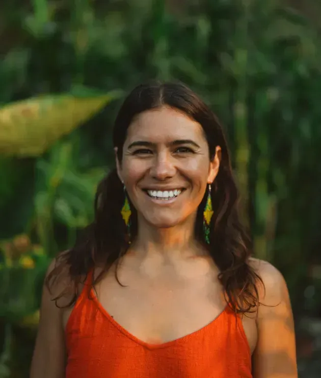 Woman smiling outdoors, wearing orange top, green leafy background, dangling earrings.