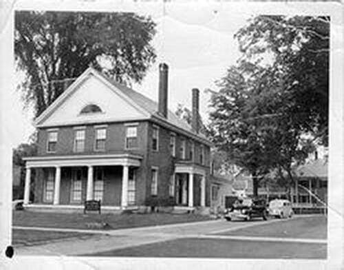 Black-and-white street view of a two-story brick house with a porch, parked cars, and tall trees