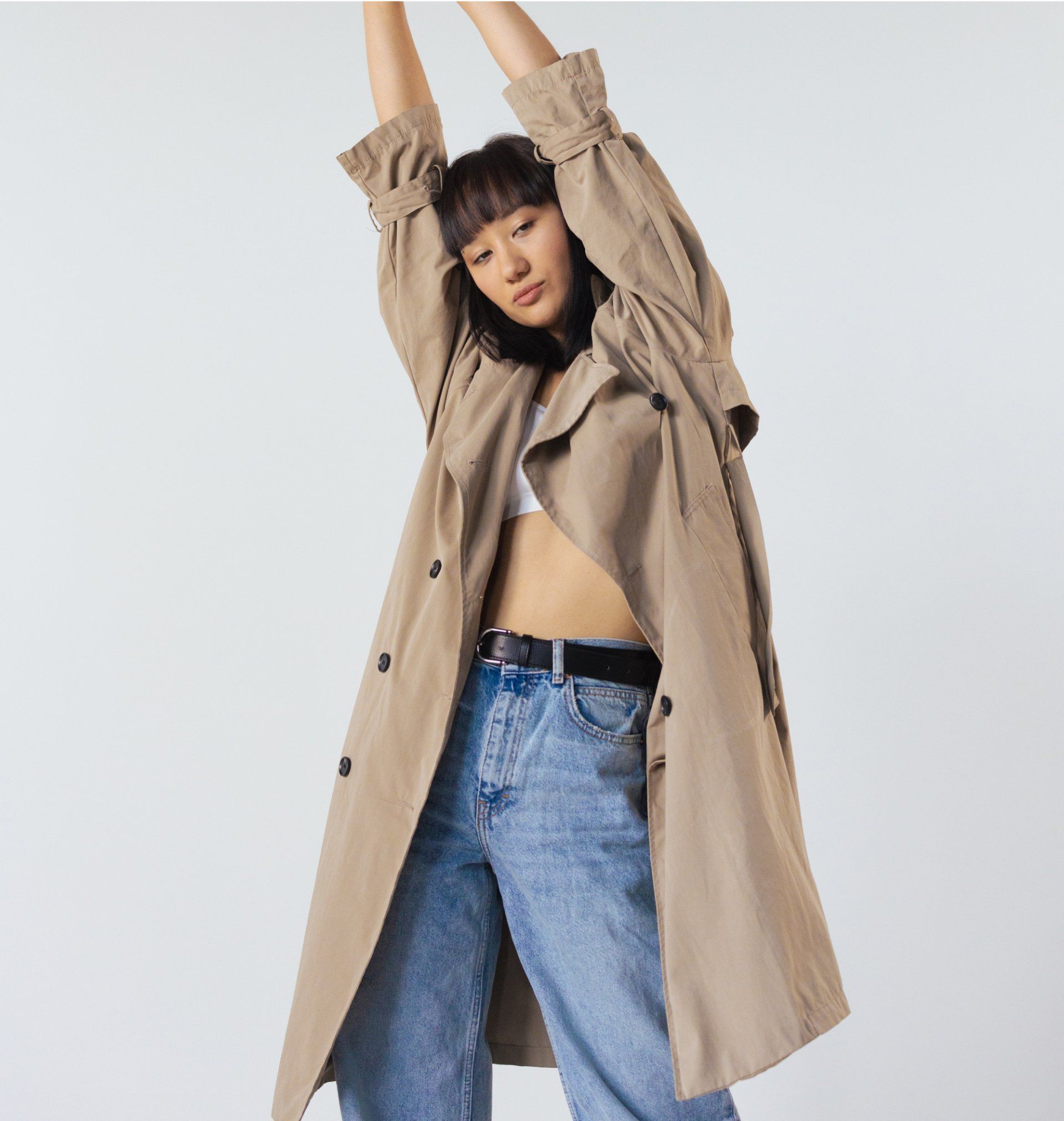 Woman in tan trench coat, looking down, hand in pocket, against a light background.