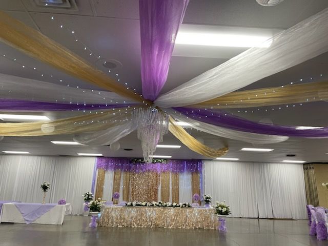 Event hall decorated with purple, gold, and white draping from the ceiling; a stage is in the back.
