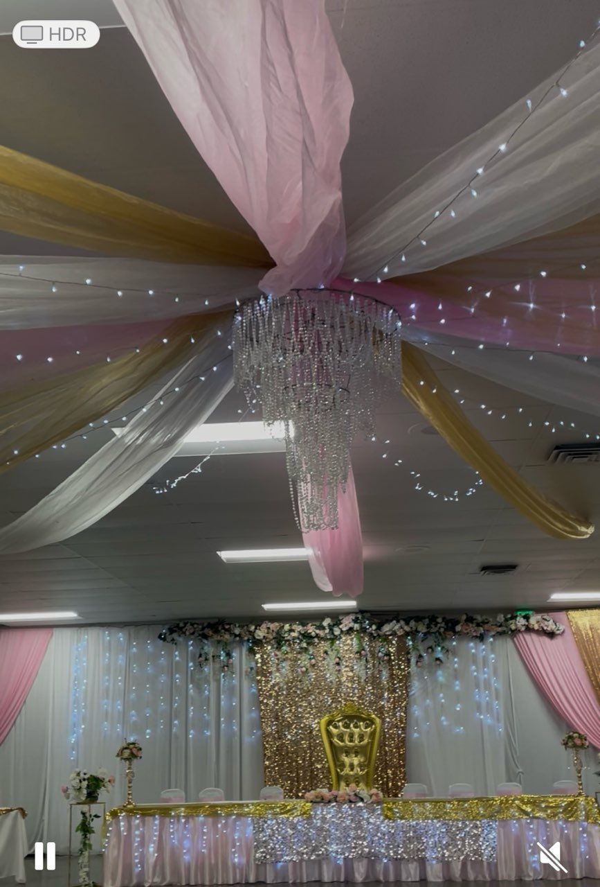Event hall decorated with pink, gold, and white draping, fairy lights, and a chandelier; head table in focus.