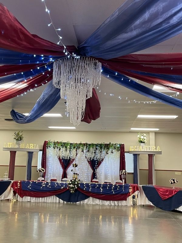 Decorated event hall with red, white, and blue draping and a chandelier. A table is set with floral arrangements.