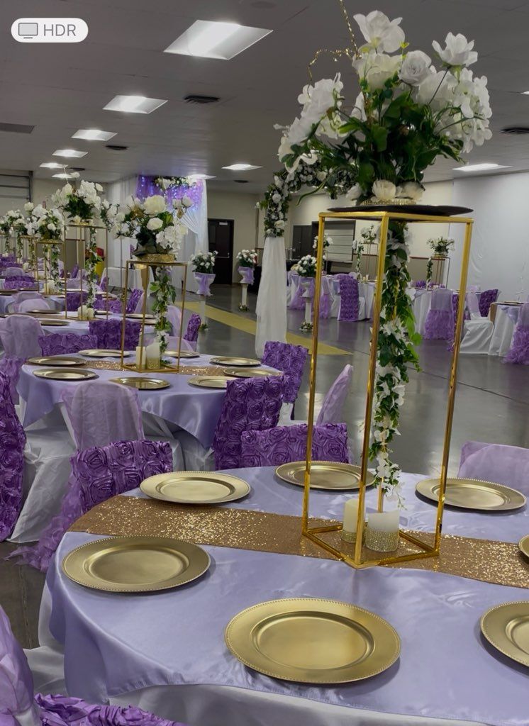 Elegant reception hall with round tables set for a celebration. Lavender tablecloths with gold chargers, floral centerpieces.