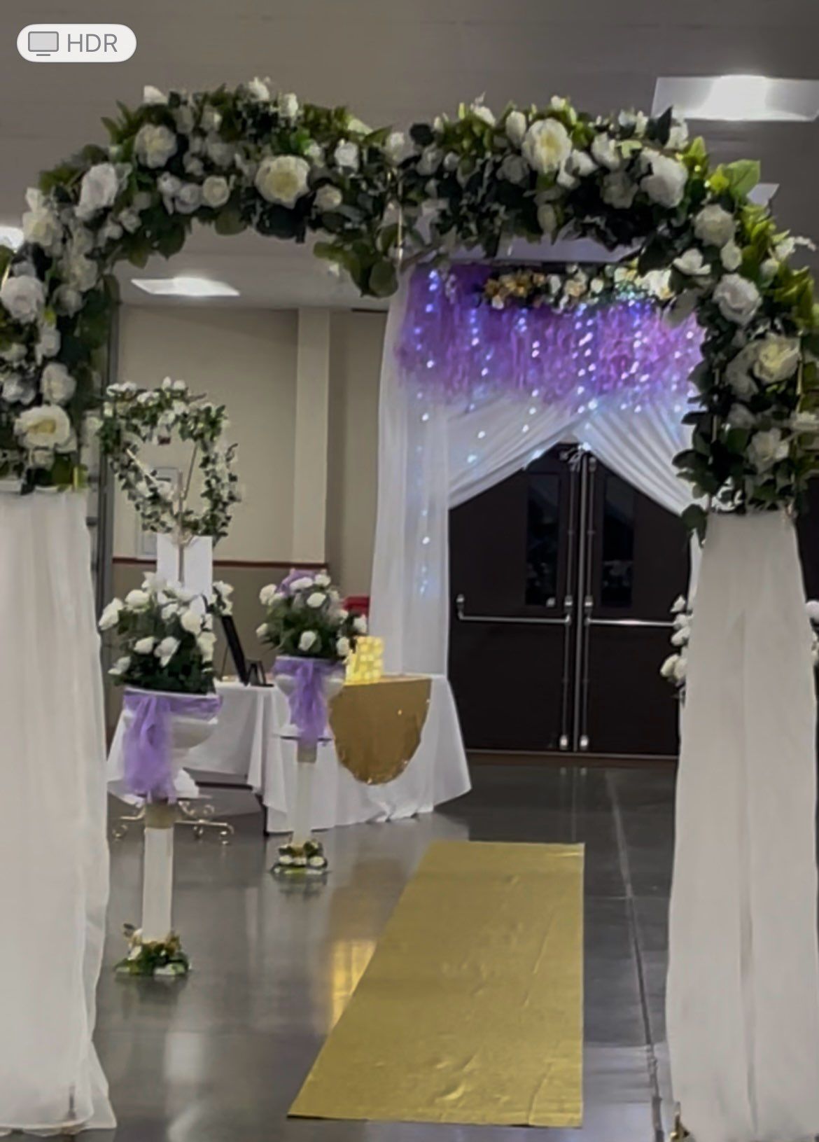 Wedding ceremony setup with white and purple flowers, gold aisle runner, and draped fabric.
