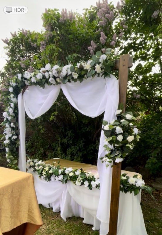 Wedding altar with white flowers, draped fabric, and gold table cloth, outdoors.