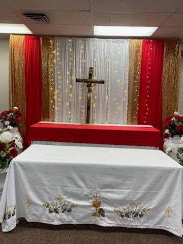 Altar with cross, draped in red, gold, and white curtains, with a white table and embroidered flowers.