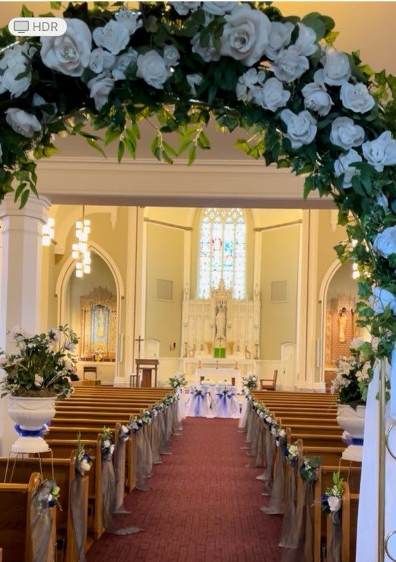 Church interior decorated for a wedding with an arch of white roses and greenery, aisle runner, and floral pew decorations.