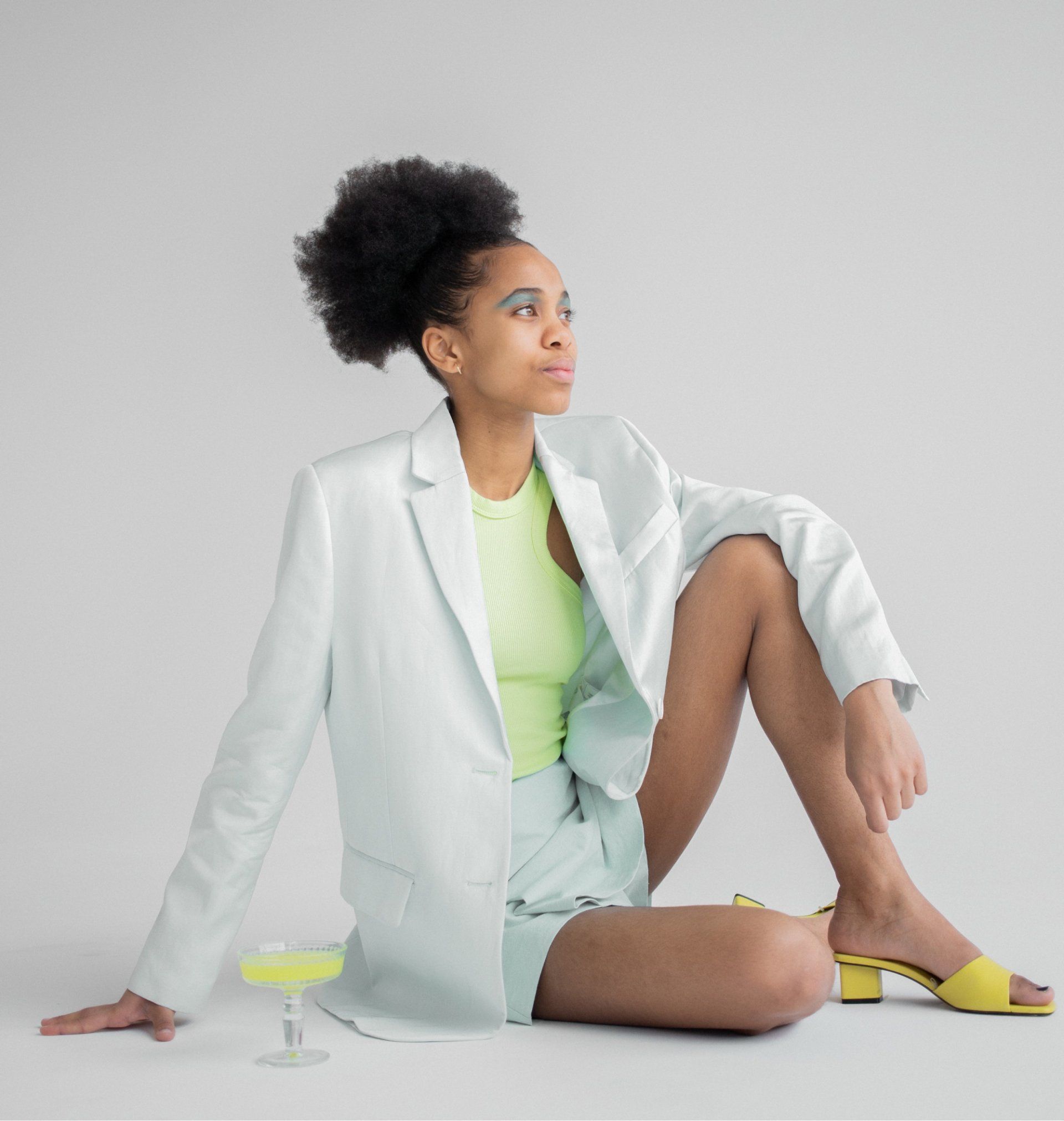 Woman in a mint green suit and neon top sits with a cocktail, looking off-camera.