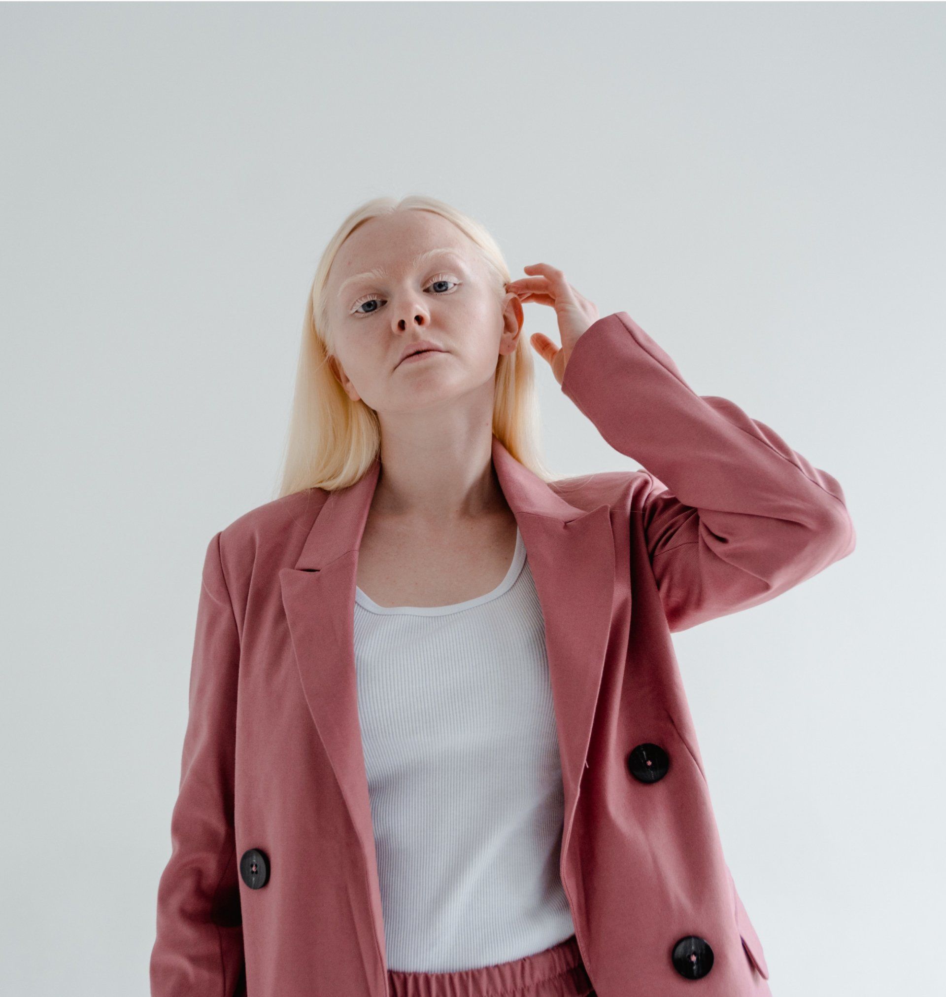 Woman with blonde hair wearing a pink blazer, white top, and pants, touching her head. White background.