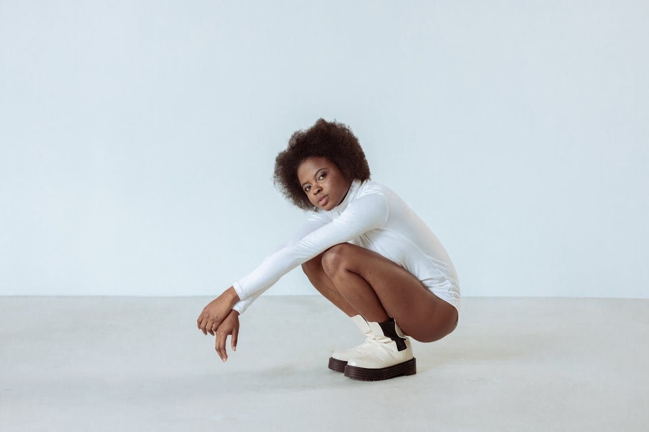 Woman in white bodysuit and boots squats on a light gray floor, arms extended, looking at the camera.