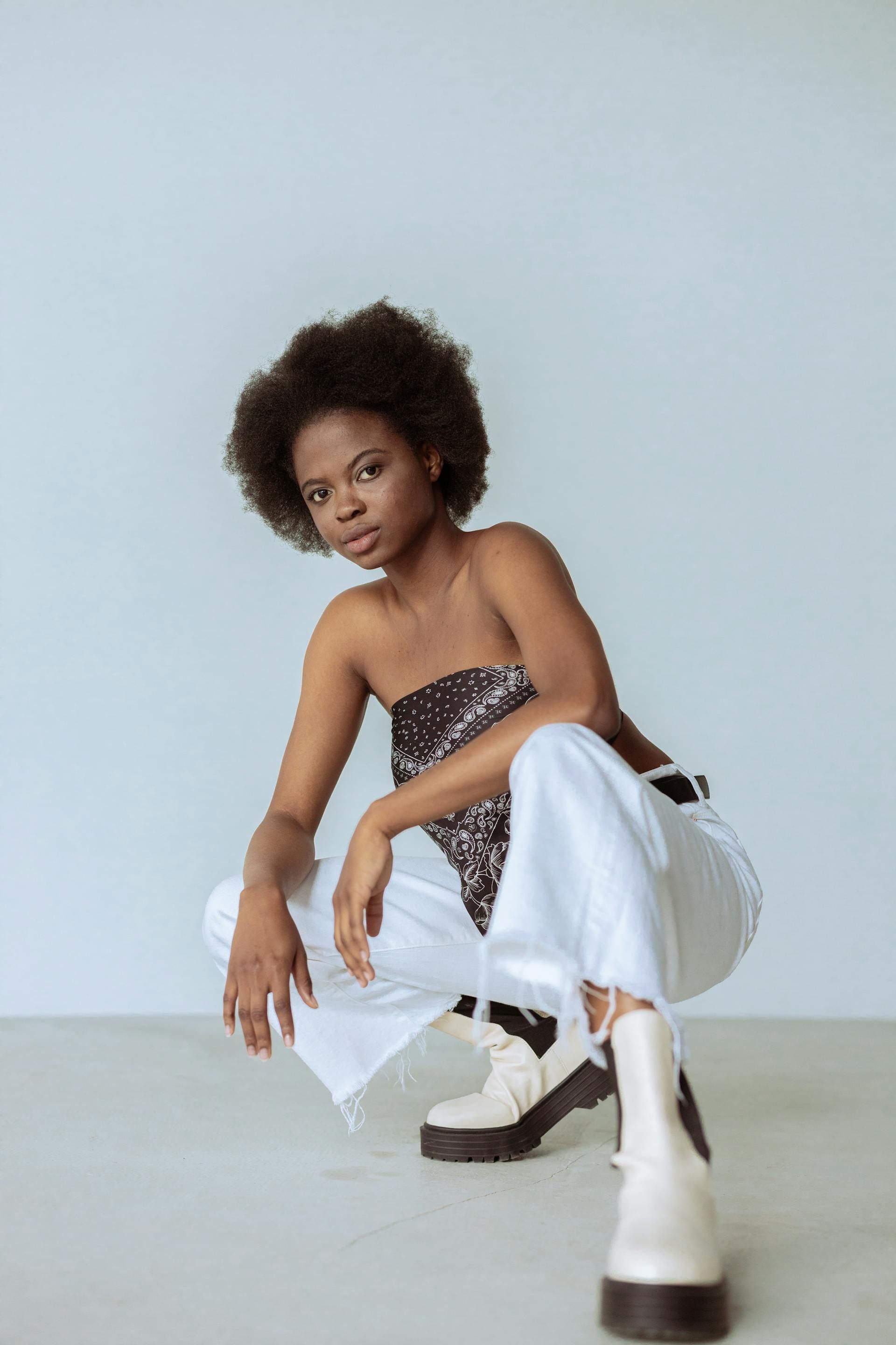 Woman in sparkly top and white pants squats, looking at the camera. White boots. Light blue backdrop.