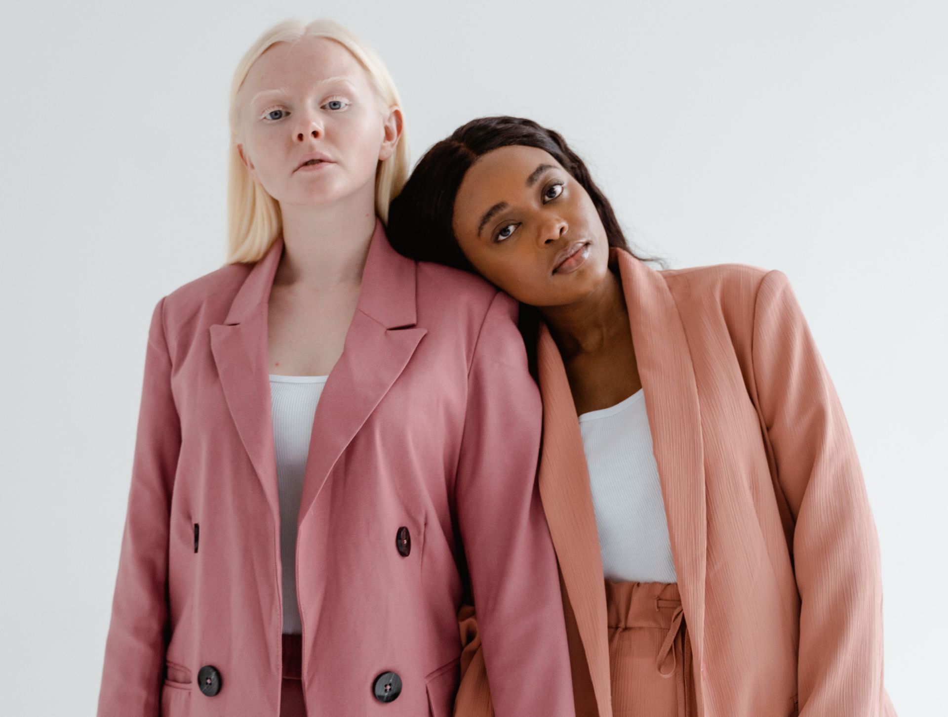 Two women in pink suits, one with blonde hair, the other with black hair, leaning on each other.