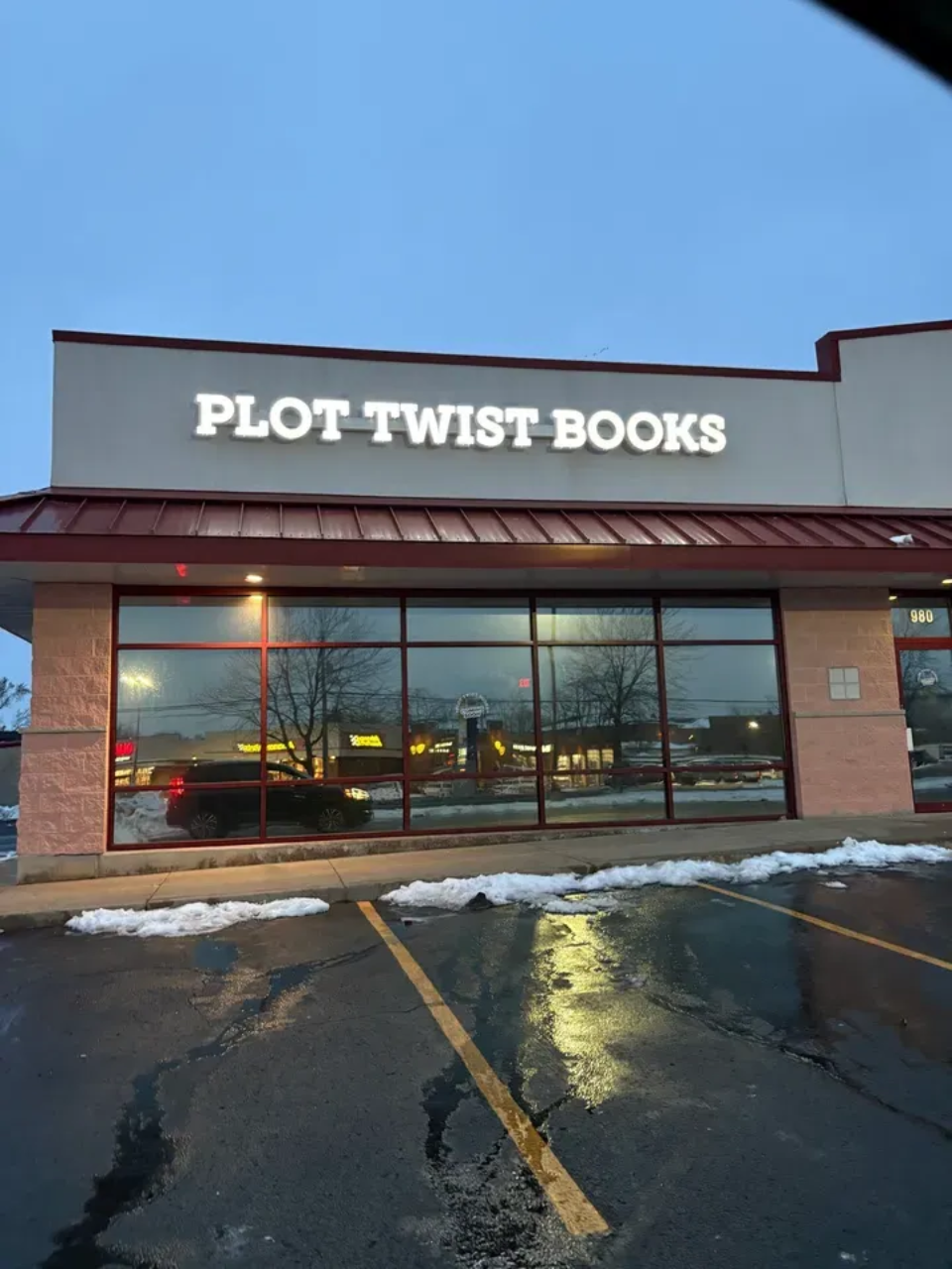 Plot Twist Books storefront with large windows, snowy curb, and wet parking lot at dusk