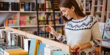 Woman browsing books in a library, reaching for a book with a blue-and-white patterned sweater.