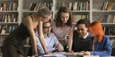 Five people gathered around a laptop in a library, collaborating and looking at the screen