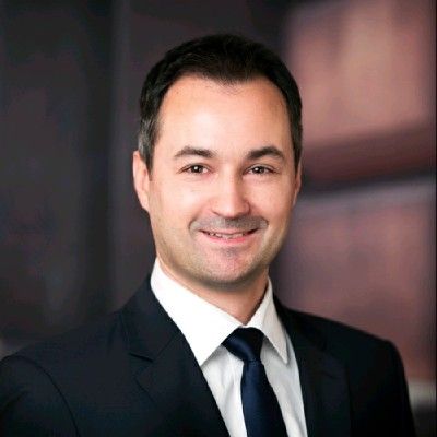 Man in dark suit, smiling, looking at the camera. White shirt and blue tie. Blurred background.