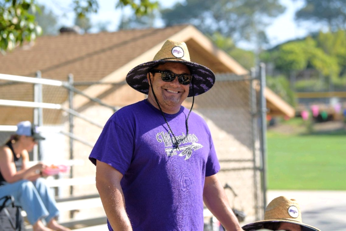 Football coach with arms crossed, looking stern on a sunny field, players in the background.