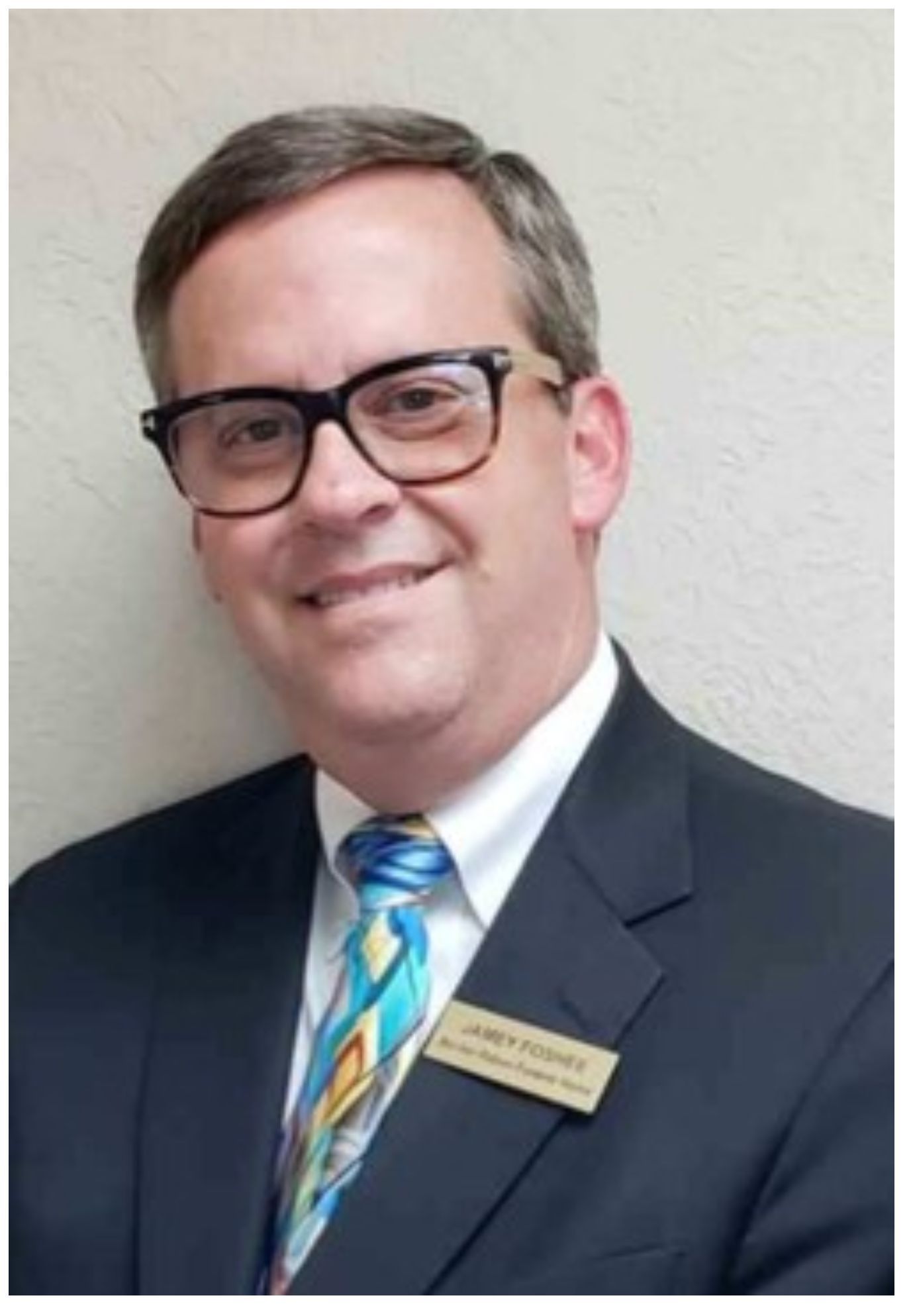 Smiling man in a dark suit, striped blue tie, and glasses, standing against a light wall.