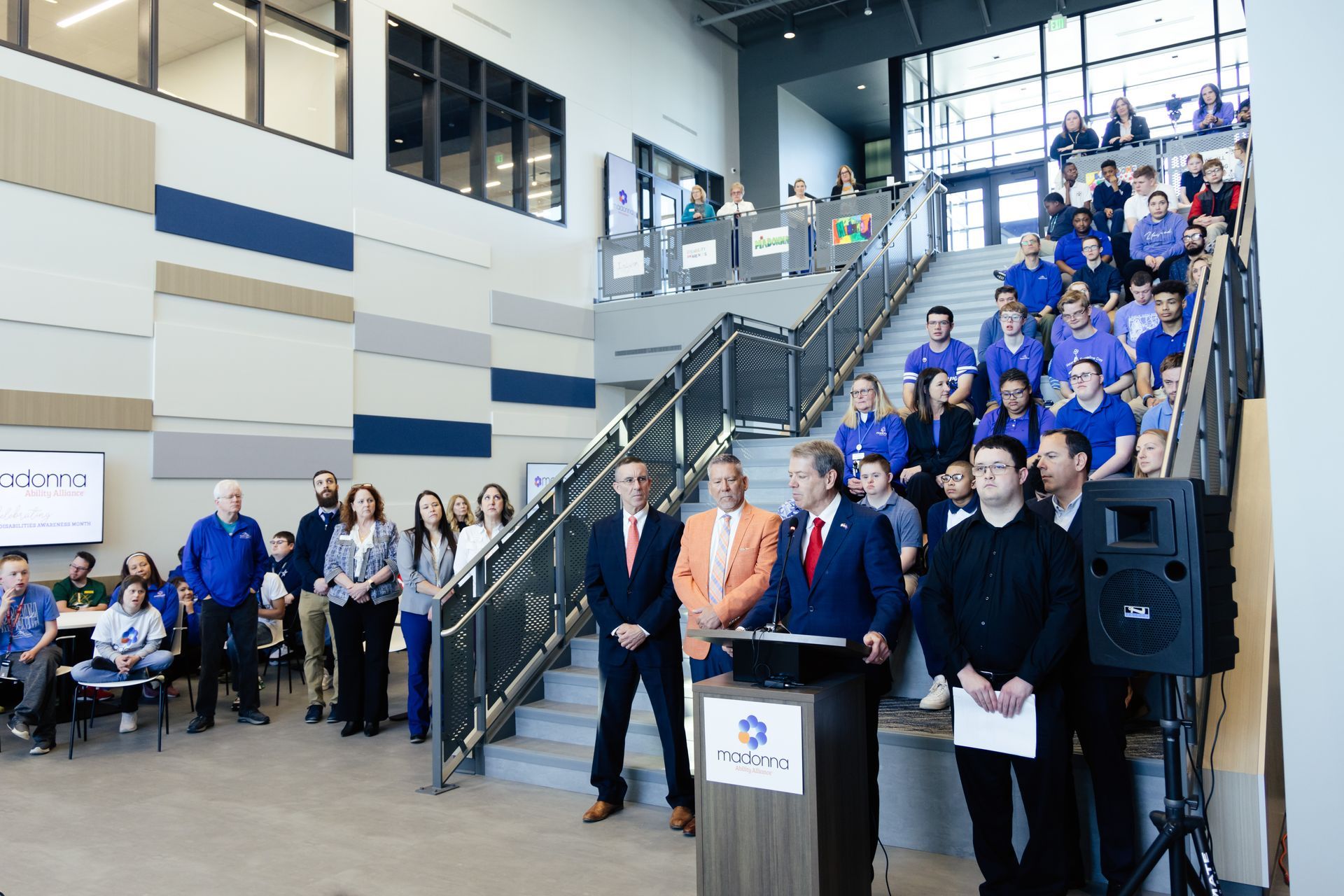 People at a podium with a blue banner, stairs filled with people, and a modern building interior.