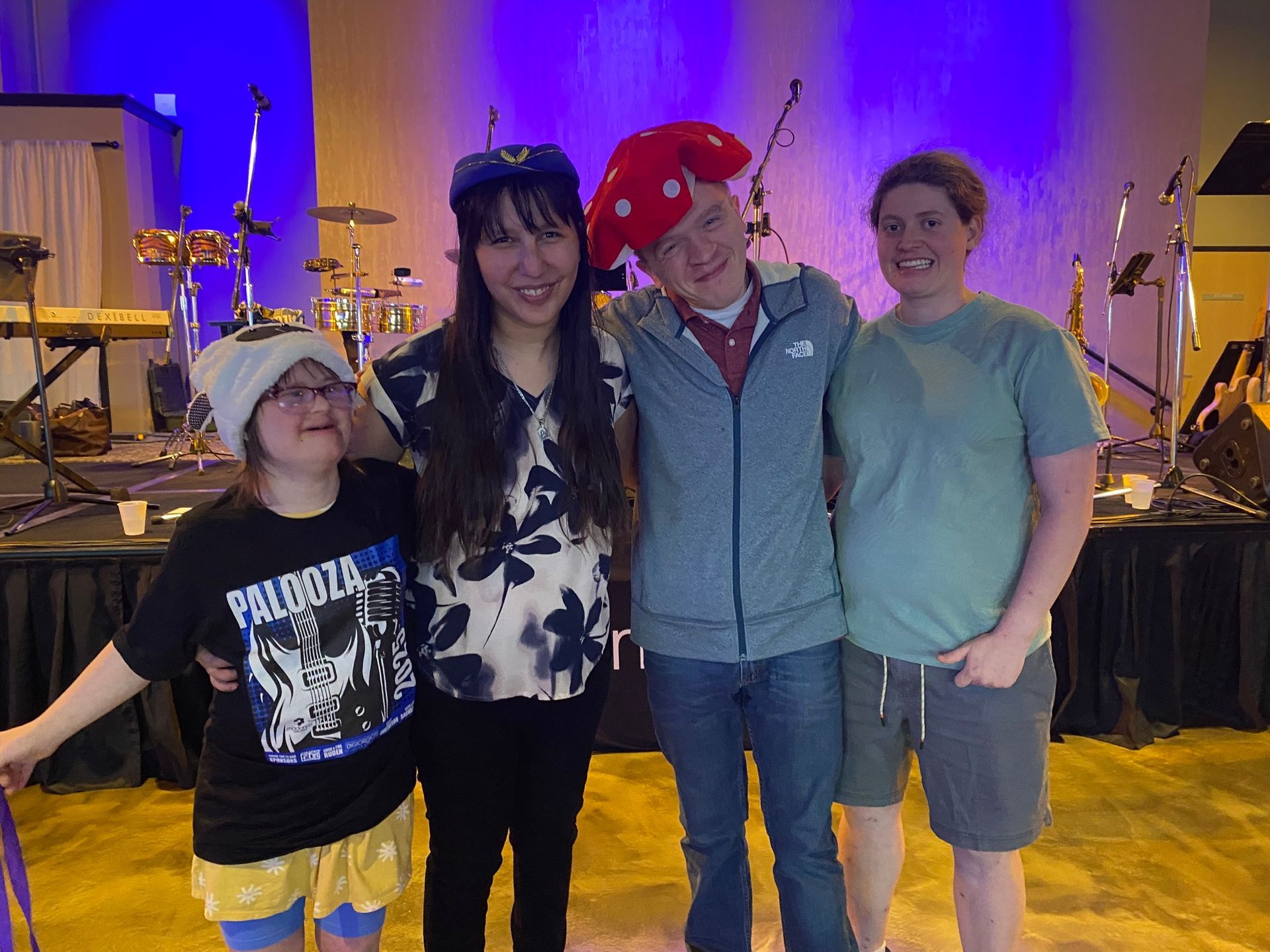 Four people in a room with a stage. Two wear hats, one a mushroom, and they are smiling while posing for the camera.