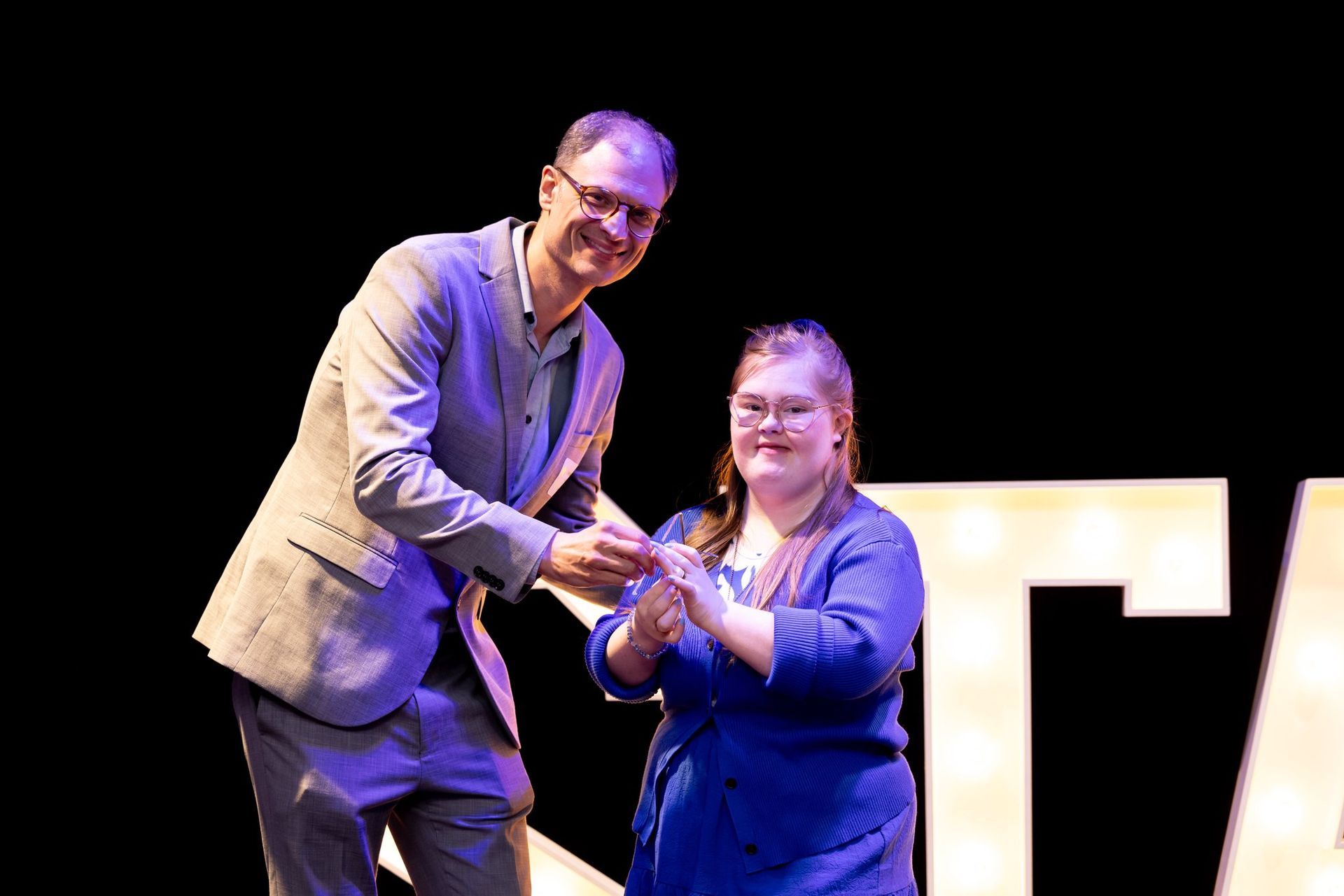 Man in blazer gives award to person in blue dress on stage. Large white