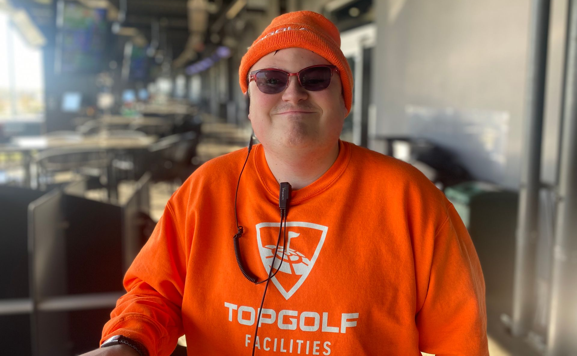 Person wearing orange Topgolf apparel and sunglasses at a Topgolf facility.