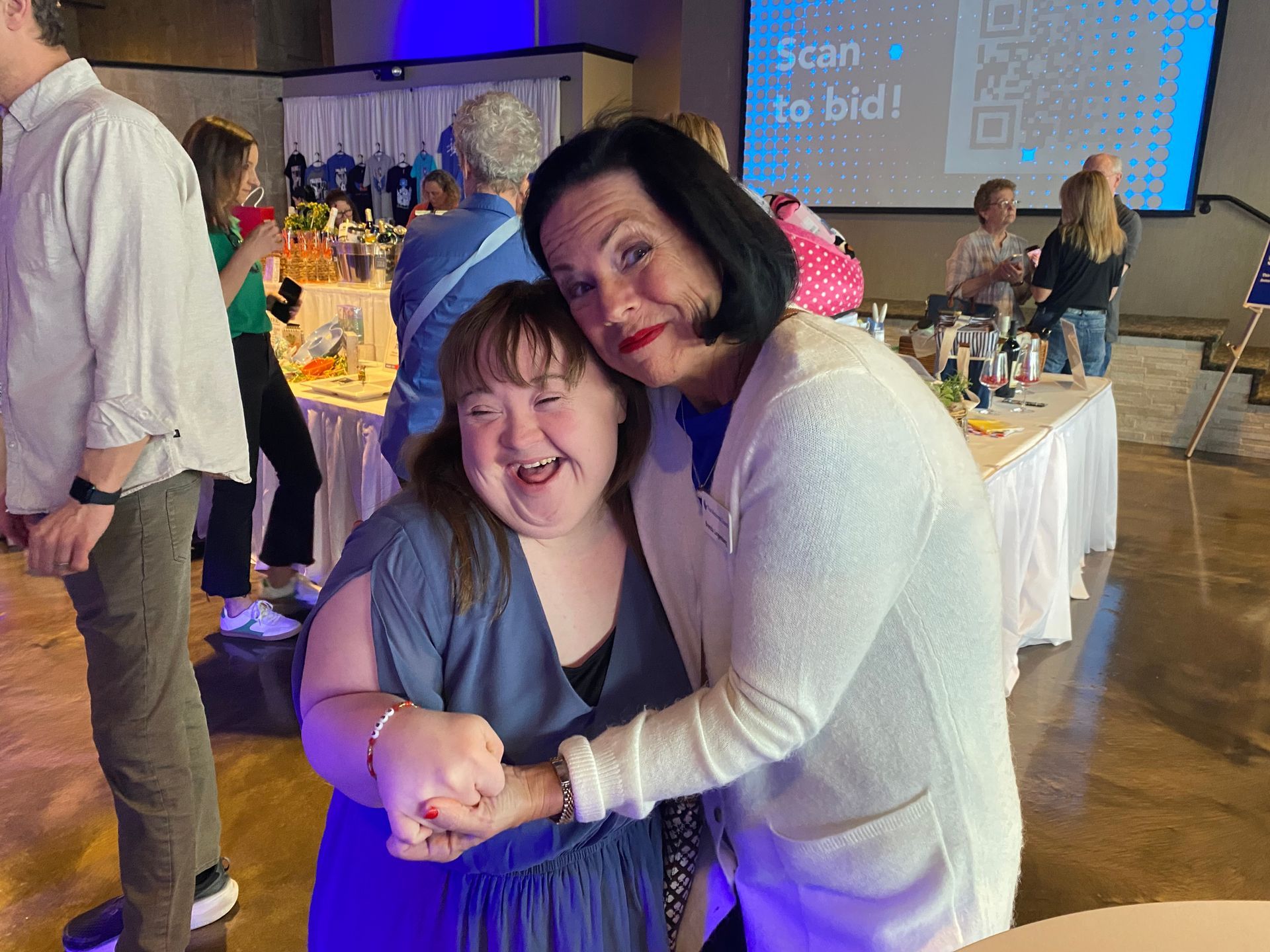 Two women smiling and embracing at an event, surrounded by tables with items. A banner reads, 