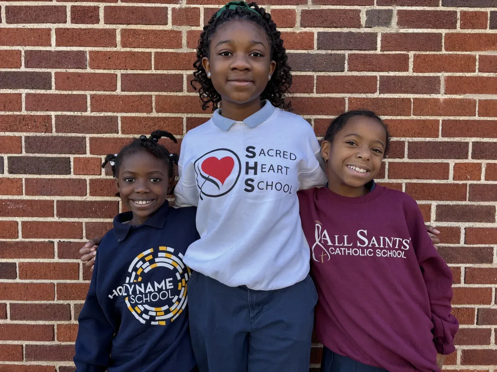 Three children smiling in front of a brick wall, wearing school uniforms: white, navy, and maroon.
