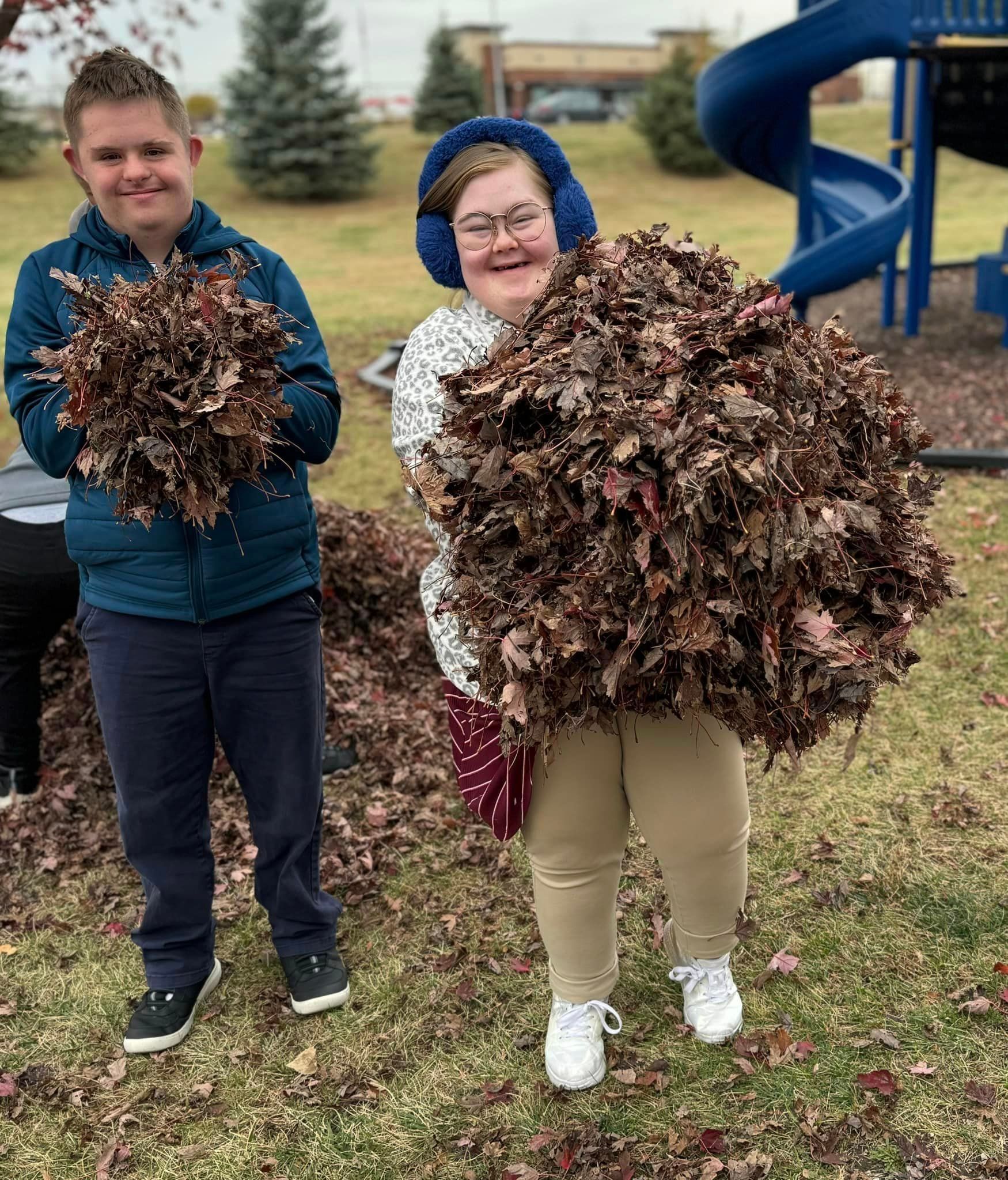 Two people holding large piles of brown leaves on a grassy playground. One wears earmuffs.