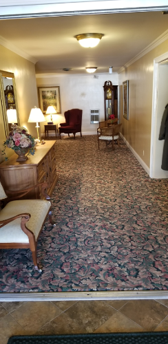 Long carpeted hallway with lamps, chairs, and a small table under warm ceiling lights
