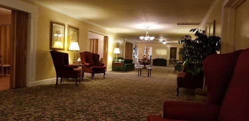 Warm hotel lounge with patterned carpet, armchairs, lamps, and ceiling lights in a softly lit hallway-like space