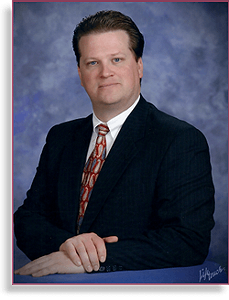 Studio portrait of a man in a dark suit and patterned tie against a blue backdrop