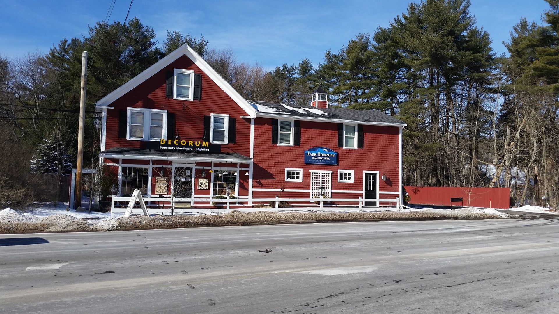 Red building with a store, snow-covered ground, and tall pine trees under a clear blue sky.