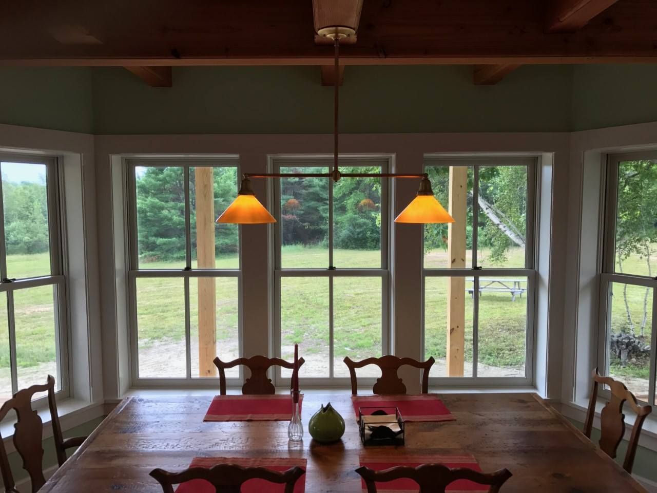 Dining room with wooden table, chairs, and windows overlooking a green field. A hanging light fixture illuminates the table.