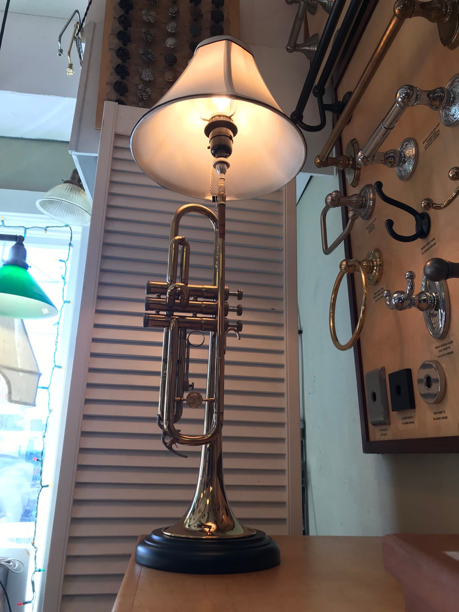 Trumpet lamp with lampshade on a shelf, near a white shuttered wall.