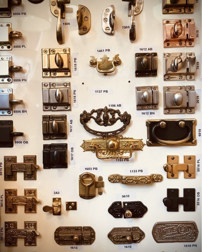 Assortment of antique cabinet latches, handles, and pulls in various metals displayed on a wall.