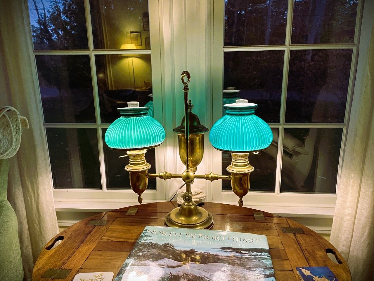 Antique brass lamp with teal glass shades on a wooden table in front of a window.